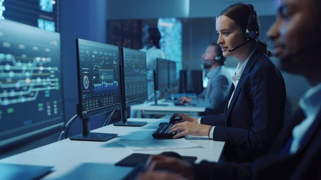 Happy Confident Technical Customer Support Specialist Having a Headset Call while Working on a Computer in a Dark Monitoring and Control Room Filled with Colleagues and Display Screens.