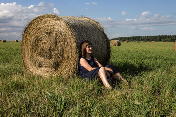 Naklejka na ściany i meble A beautiful girl sits on a field in beautiful summer weather near a haystack. The girl dreams. The joys of rural life. Man and nature. Peace and grace. Escape from civilization.