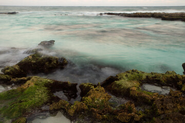 Long exposure shot of the beach at sunset. Surreal water smoky effect. View of the shore mossy rocks, turquoise ocean water and blurred sea waves with a beautiful dusk light.