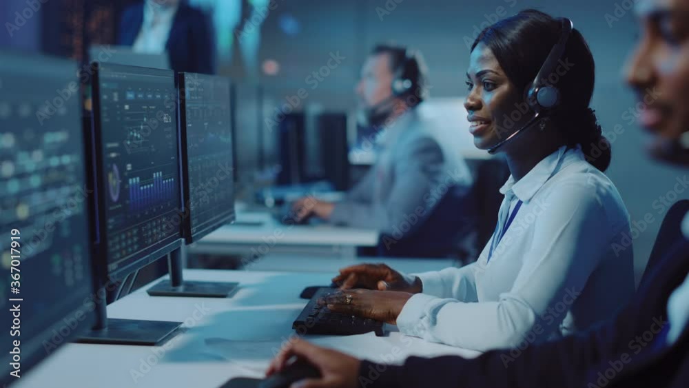 Leinwandbilder Pretty Black Technical Customer Support Specialist is Talking on a Headset while Working on a Computer in aCall Center Monitoring and Control Room Filled with Colleagues and Display Screens.