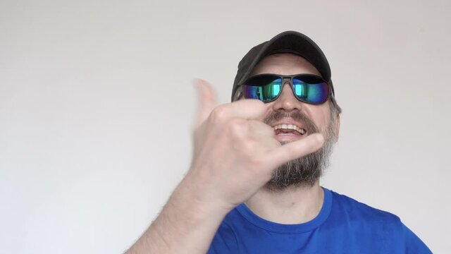 Adult man in blue t-shirt and sunglasses shows gesture with his hand and smiles.