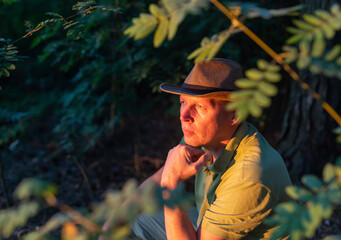 American Traveler mature man in cowboy hat is looking at sunset sitting in forest behind trees and...