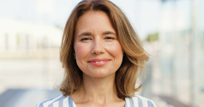 Portrait of beautiful Caucasian woman with fair hair looking at side, turning face to camera and smiling outdoors on sunny summer day. Happy female at street. Close up.