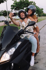 Fototapeta na wymiar Cheerful Stylish Couple Satting On Modern Motorbike Outdoors and making a selfie with a smartphone