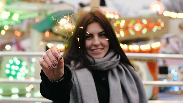 Portrait of young smiling woman holding sparklers and laughing in the evening city, amusement park slow motion