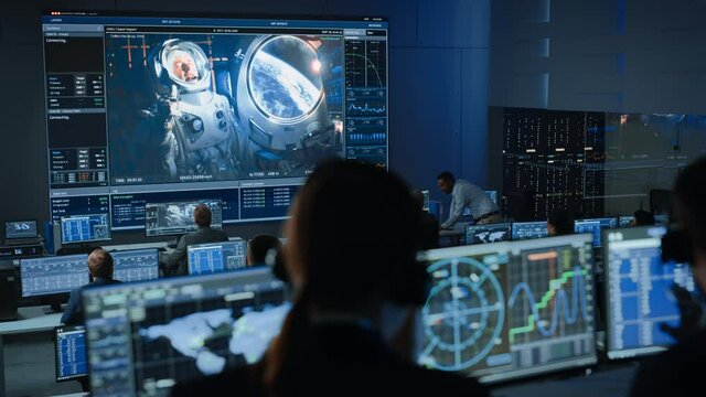 Group of People in Mission Control Center Establish Successful Video Connection on a Big Screen with an Astronaut on Board of a Space Station. Flight Control Scientists Sit in Front of Computers.