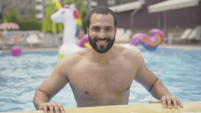 Portrait of handsome young Middle Eastern man posing in pool at luxurious summer resort. Bearded athletic male tourist with toothy smile standing at poolside and looking at camera.