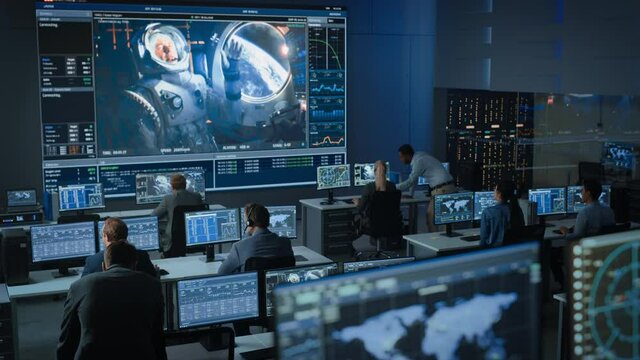 Group of People in Mission Control Center Establish Successful Video Connection on a Big Screen with an Astronaut on Board of a Space Station. Flight Control Scientists Sit in Front of Computers.