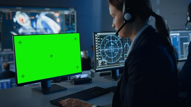 Green Screen Horizontal Mock Up on a Computer Display Used by a Female Controller in a Mission Control Center Room with Other Scientists. Team of Engineers Work in Monitoring Room Full of Displays.