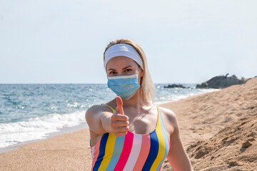 beautiful girl in a medical mask on the beach shows thumbs up