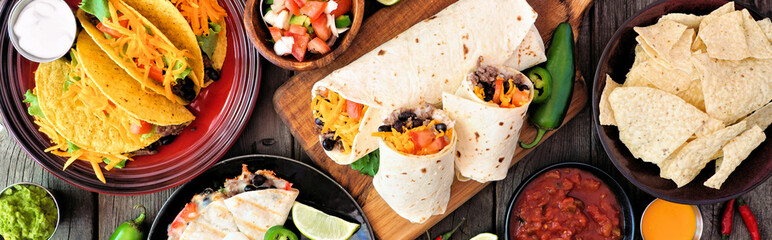 Mexican food table scene banner. Top down view over a dark wood background. Tacos, burritos,...