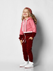 Little blonde curly beautiful girl in red sport costume and white sneakers standing with hand in pocket and laughing