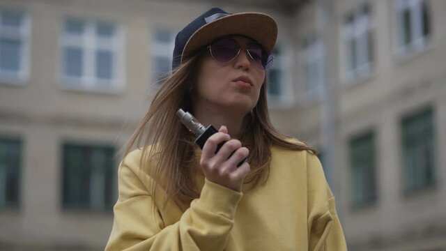 Pretty young hipster woman in black cap yellow hoodie vape ecig. Girl vape popular ecig gadget, vaping device.Happy brunette vaper girl with e-cig. Portrait of smoker female model with electronic