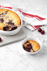 Serving of Homemade Cherry Cobbler in a White Bowl on a White Marble Countertop; Dish of Cobbler in Background; Fresh Cherries