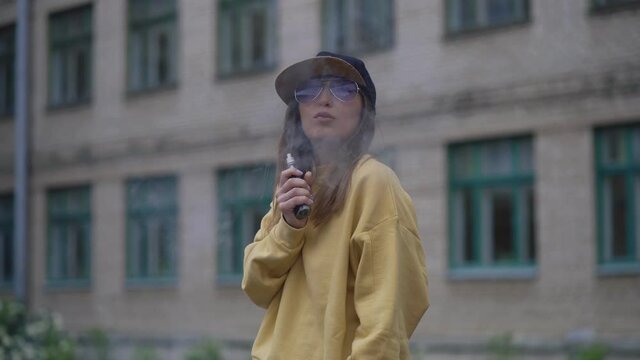 Pretty young hipster woman in black cap yellow hoodie vape ecig. Girl vape popular ecig gadget, vaping device.Happy brunette vaper girl with e-cig. Portrait of smoker female model with electronic