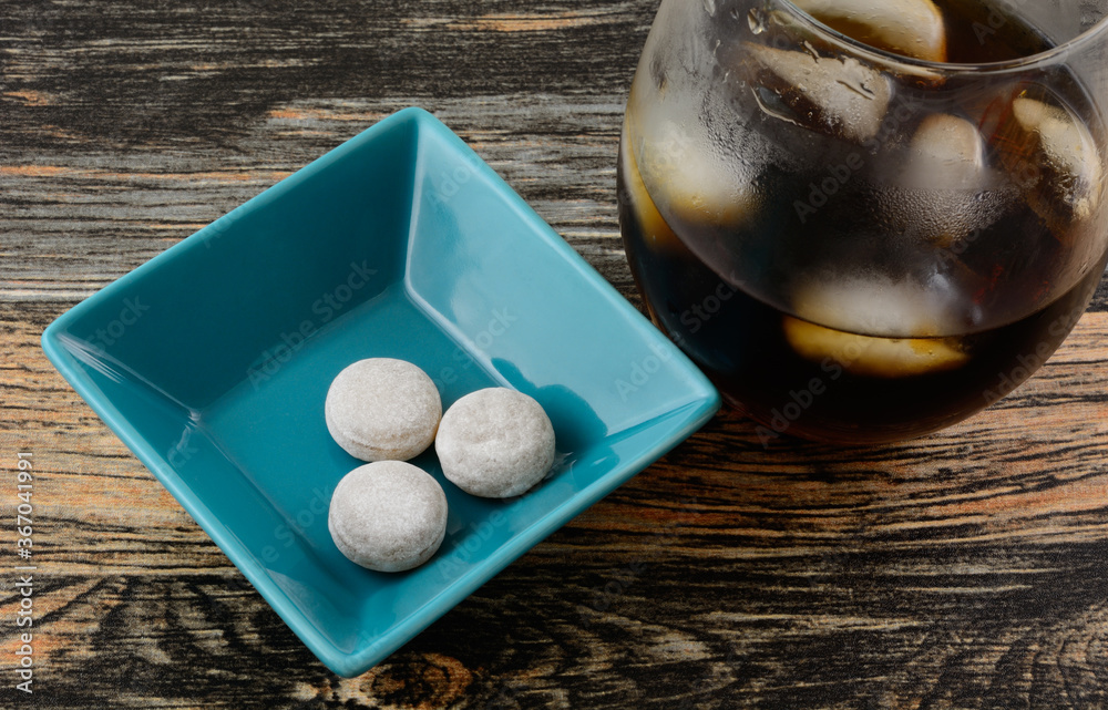 Poster cola flavored hard candy in blue candy dish with glass of cola with ice cubes on side