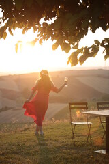 woman in a burgundy dress drinks red wine from a glass at sunset in the mountains.