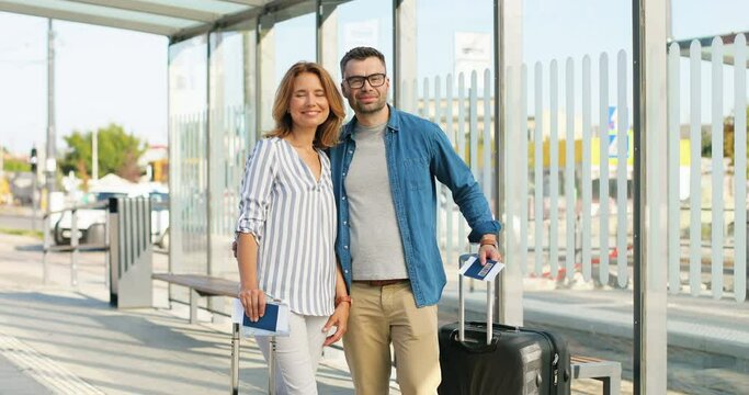Portrait of young Caucasian happy married couple standing at train station with suitcase on wheels and smiling to camera on sunny summer day. Beautiful woman and handsome man tourists. Travelling Trip