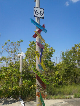 Pole with Route 66 highway shield at top and place names below on colored signs.