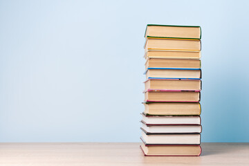 A tall stack of books stands on a wooden table against a light blue wall, space for text