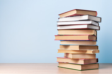 A tall, chaotic stack of books stands on a wooden table against a light blue wall, space for text
