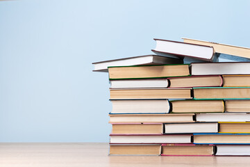 A large stack of books stands on a wooden table against a light blue wall, space for text