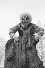 Halloween holiday concept. Creepy clown costume. Spooky clown near old rotten stump on blurry foggy trees background.Black and white halloween photo in retro style.Autumn holidays.Horror and fear.