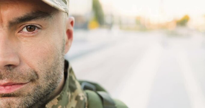 Portrait of Caucasian handsome young man soldier in cap with backpack looking confident at street. Close up of half face of male militarian with wise brave face outdoor at bus stop. Military uniform.