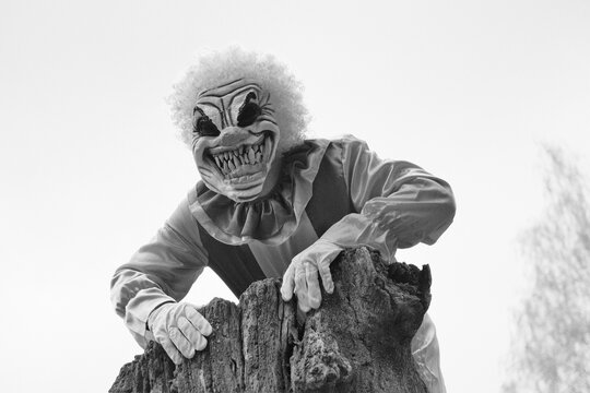 Halloween. Creepy clown costume. Spooky clown near old rotten stump on blurry foggy trees background.Black and white halloween photo in retro style.Autumn holidays.Horror and fear.