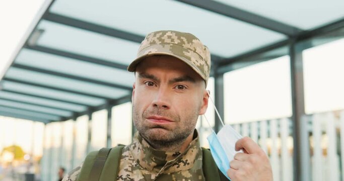Portrait of happy Caucasian handsome young male soldier in cap taking off medical mask at street. Close up of confident man militarian outdoor in town during pandemic. Military uniform.