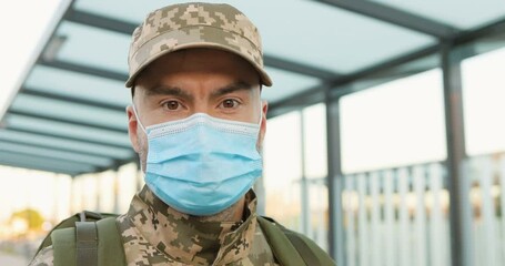 Portrait of serious Caucasian handsome young male soldier in cap and medical mask standing at street. Man militarian looking at camera at train station. Bus stop. Military uniform. Covid-19 pandemic. - Powered by Adobe