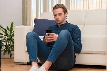 young man sitting on the ground and leaning on a sofa focused on his phone. Communication concept.