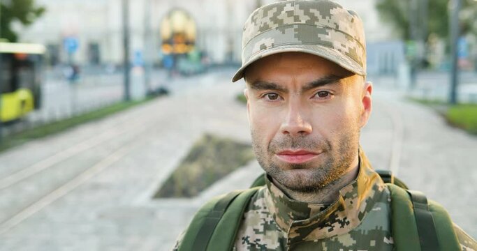 Portrait of happy Caucasian handsome young male soldier in cap with backpack looking brave at street. Close up of confident man militarian outdoor in town. Military uniform.