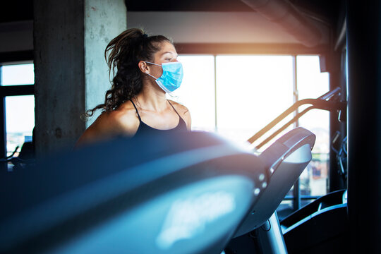 Woman training in gym during covid-19 pandemic and coronavirus. Sportswoman running on treadmill and wearing hygienic face mask to protect herself against highly contagious corona virus.