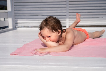 A little girl lies on a pink shawl on a white background, sunbathes. The eyes look slyly to the side. The right leg is bent at the knee, the heel is visible. Girl in an orange swimsuit. High quality