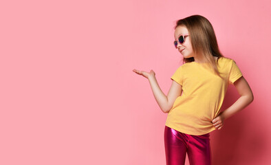 Cute frolic kid girl in yellow t-shirt, shiny legging and sunglasses holds hand up looks at open palm. Free copy space