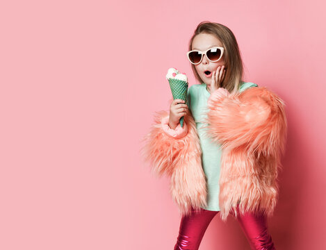 Kid Girl In Faux Fur Coat And Glossy Leggings Is Surprised At How Big Ice Cream With Candies In Waffles Cone She Holds Is