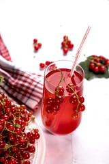 Sprigs of red currant berries are lying on a white plate, a berry Morse stands in a glass glass on a light background, next to a napkin in a red and white check. Summer drink that quenches your thirst