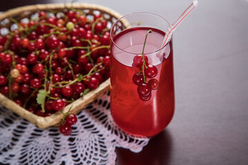Sprigs of redcurrant berries lie on a wicker plate, and a Morse of berries stands in a glass glass and a tube on a dark table with a knitted napkin. A summer drink that quenches your thirst.