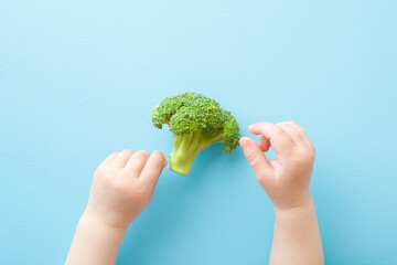 Naklejka na ściany i meble Baby hands touching piece of green broccoli on light blue table background. Pastel color. Fresh vegetable. Closeup. Point of view shot. Top down view.