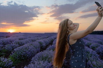Smiling beautiful woman with long hair taking selfie at blooming blossoming beautiful landscape of violet purple lavender flowers on field with summer sunset and orange sky, Bulgaria. Beauty concept.