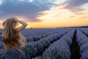Beautiful girl with hands up and long hair stays with back near incredible landscape of violet purple lavender flowers on field with summer sunset and orange sky, Bulgaria. Beauty production concept.