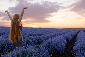 Beautiful girl with hands up and long hair stays with back near incredible landscape of violet purple lavender flowers on field with summer sunset and orange sky, Bulgaria. Beauty production concept.