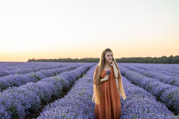 Beautiful romantic girl with long hair and in orange dress holds a bunch of lavender flowers and enjoying their fragrance among blooming violet purple lavender field at summer sunset. Travel concept.