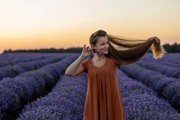 Pretty happy girl in orange dress with long hair holds a lavender bouquet in blooming blossoming beautiful landscape of violet purple lilac lavender field with sunset and orange sky. Summer concept.