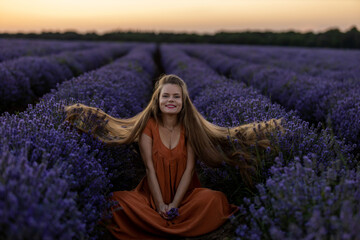 Smilig happy girl with long hair on lavender bushes and in orange dress sits in an incredible violet purple lavender field at sunset and orange sky, Woman holds blooming flower. Summer travel concept.