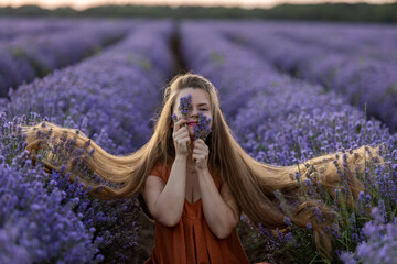 Smiling romantic girl with long hair and in orange dress holds a bunch of lavender flowers and enjoys the fragrance. Girl sits in an incredible violet lavender field at sunset. Summer travel concept.