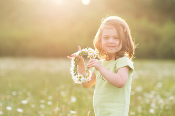 Cute baby girl 2-3 year old playing with chamomile wreath in meadow over sunny background closeup. Looking at camera. Childhood.