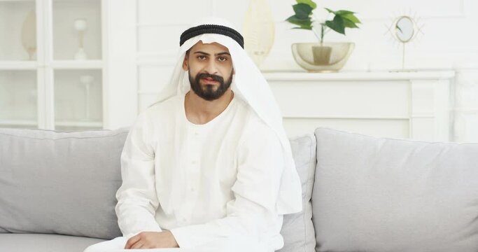 Portrait of handsome young Arabian man in kandura sitting on couch in living room and smiling to camera. Male Arab muslim in dishdasha on sofa at home. White interior Arabic millioner from Middle East