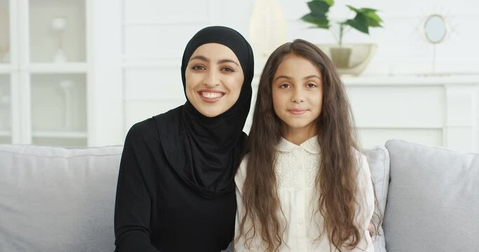 Portrait of beautiful young muslim mother in headscarf smiling to camera while sitting with cute teen daughter on sofa in living room. Pretty Arabian woman in hijab and small teenage girl on couch.